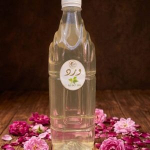 Bouteille transparente d'eau de rose artisanale posée verticalement sur une table en bois rustique, entièrement entourée de roses roses entières et de pétales éparpillés. L'étiquette ronde "ورد" est visible sous un éclairage chaleureux.